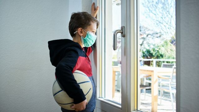 Niño con mascarilla y una pelota bajo el brazo mirando hacia afuera sin poder salir a jugarG