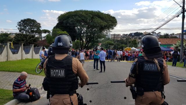 Dois PMs fazem segurança de área da PF em Curitiba; ao fundo, uma fita amarela delimita a área de segurança; atrás da fita, há manifestantes
