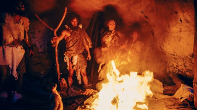 Tribo de caçadores-coletores pré-históricos vestindo peles de animais ao redor da fogueira do lado de fora de caverna à noite. Retrato de família neandertal/homo sapiens fazendo ritual de religião pagã perto do fogo