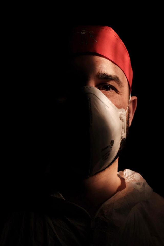 A male nurse wearing a face mask with a dark background behind him.
