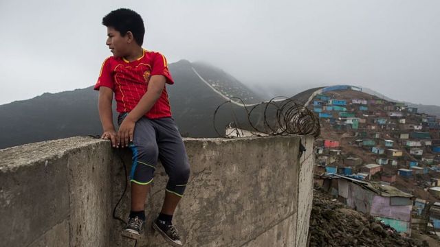 Un niño camina cerca del muro del lado donde se levantan casas humildes.