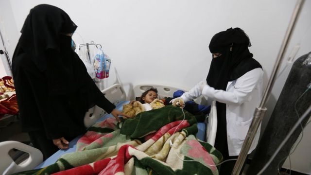 Uma criança internada com sarampo no Iêmen aparece em maca, observada por duas mulheres