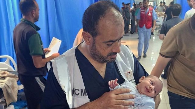 Dr. Muhammad Abu Salamiya segurando um bebê em uma movimentada enfermaria de hospital