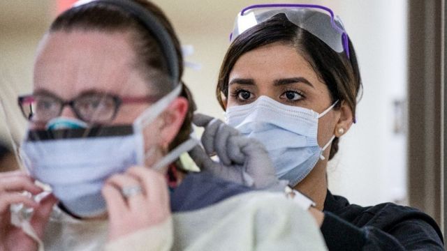 Uma profissional de saúde com máscara ajuda outra a colocar equipamento de proteção
