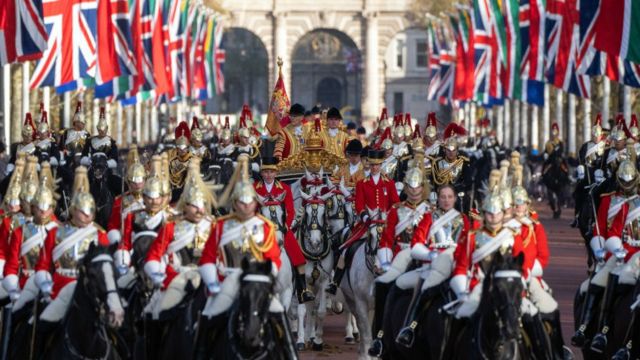 A huge amount of horses are ridden by soldiers down the Mall in London. British flags line the street on both sides. The royal carriage is in the middle.