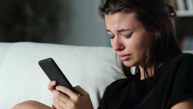 Mulher chorando no sofá, com celular na mão