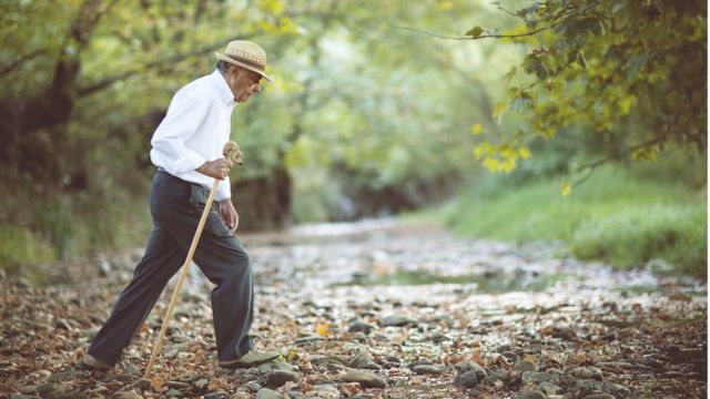 La imagen en color muestra a un anciano marrón con sombrero y ropa sosteniendo un palo y caminando por un sendero rodeado de árboles.