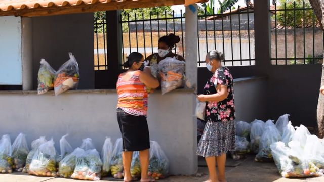 Uma mulher entrega e duas recebem cestas básicas em ação de doação promovida pelo Centro de Agricultura Alternativa do Norte de Minas