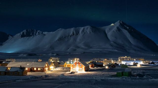 Pequeno assentamento da cidade de Ny-Ålesund — com luzes acesas enquanto a escuridão cai sobre as montanhas ao fundo