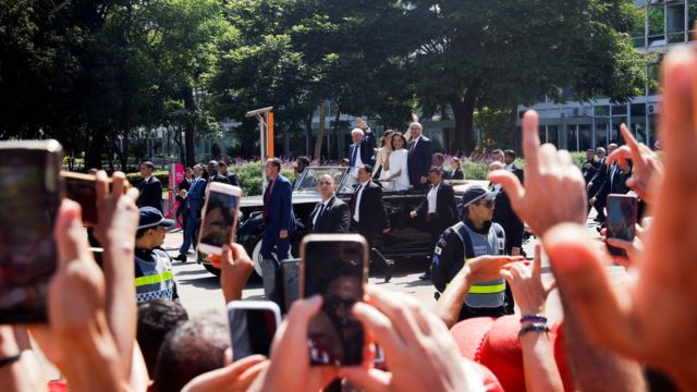 No primeiro plano, apoiadores de Lula acenando e tirando fotos; ao fundo, aparece carro conduzindo o presidente, o vice e suas esposas