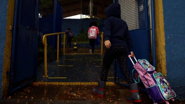 Menina de costas carrega mochila de rodinha, entrando na escola