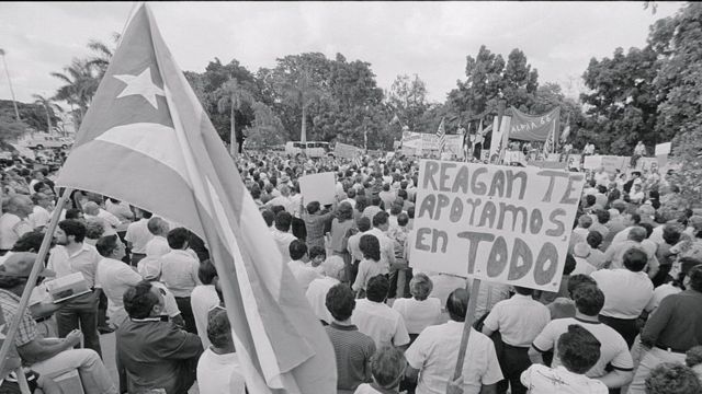 A bandeira cubana ao lado de um cartaz que diz, em espanhol, 'Reagan, te apoiamos em tudo' durante uma manifestação em 1983