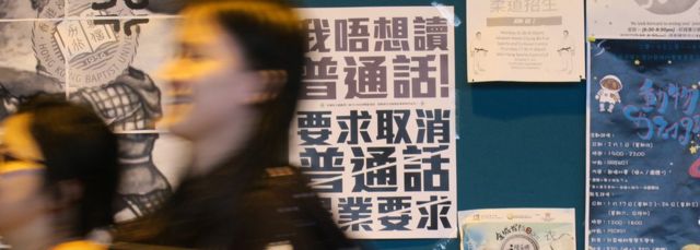 香港浸會大學逸夫校園內一群女學生走過貼上了反對普通話畢業門檻豁免試標語的學生會民主牆（BBC中文網圖片22/1/2018）