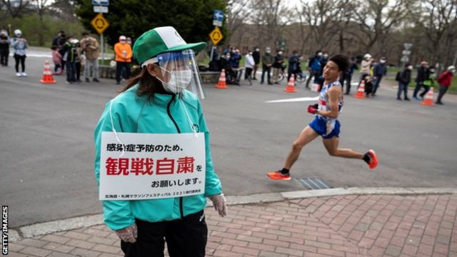A volunteer (L) holds a placard asking people to refrain from watching the competition to prevent the spread of the Covid-19 coronavirus while an athlete (R) competes in the half-marathon race which doubles as a test event for the 2020 Tokyo Olympics