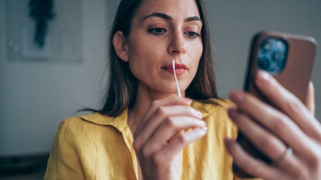 Mulher prestes a colocar swab em seu nariz olha para o telefone