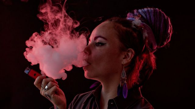 Una mujer, en semisilueta sobre un fondo negro, exhala una gran nube de vapor de cigarrillo electrónico. Su cabello está trenzado y atado por encima de su cabeza en un moño grande. Ella sostiene una pluma de vape roja en su mano visible.