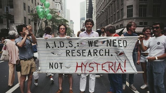 Em 1983, homens erguem um cartaz com os dizeres: 'Aids: nós precisamos de pesquisa, não de histeria'