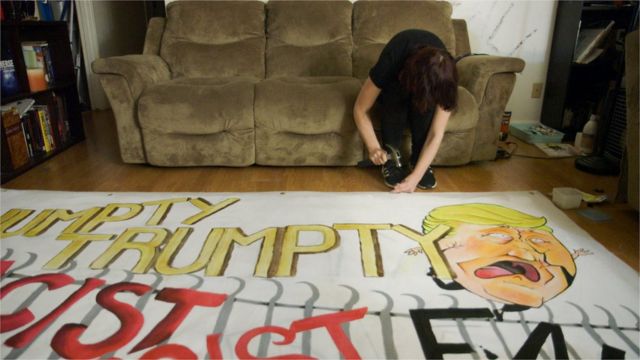 Sentada em um sofá, ativista finaliza cartaz com caricatura de Trump