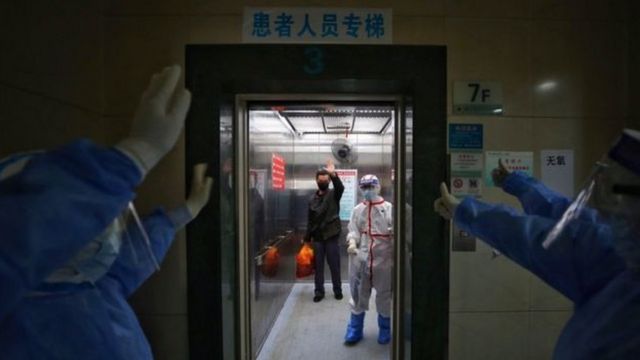 De corredor, equipe de hospital em Wuhan se despede de paciente curado da covid-19, que também acena de dentro de elevador
