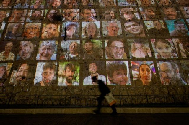 Vulto de pessoa passando, a noite, em frente a muro onde são projetadas fotos de reféns; 