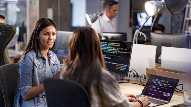 Duas mulheres sentadas conversando em frente a computadores