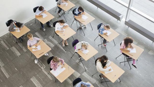 Foto tirada do alto mostra várias crianças fazendo atividade com papel em suas carteiras da escola