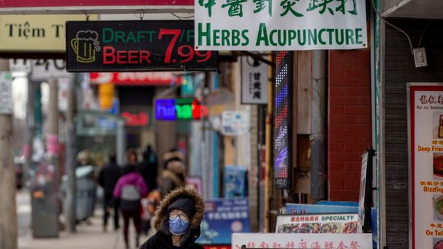 Pessoa com máscara anda em calçada de Chinatown, de onde se vê placas de comércio de cerveja e acunpuntura