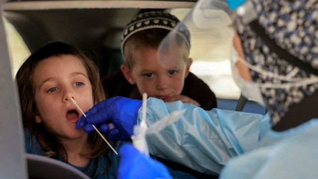 Duas crianças aparecem dentro do carro, uma delas com coleta de material do nariz para teste, feita por profissional de saúde que aparece de costas