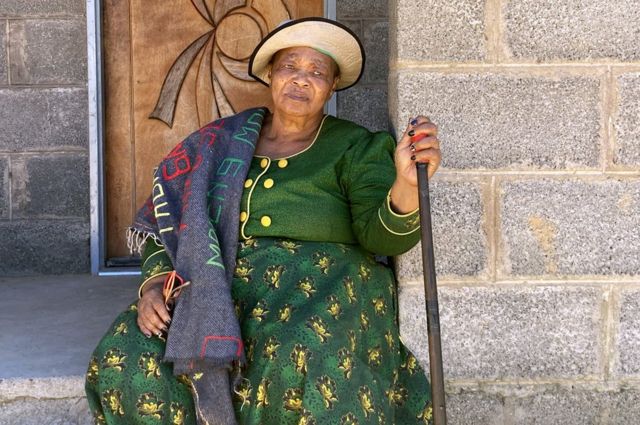 Mulher negra sentada à porta com chapéu e bengala na mão