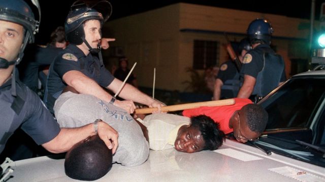 Policiais brancos empurram pessoas negras contra o capô de um carro