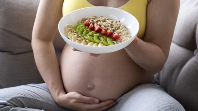 Mulher grávida comendo prato de frutas e cereal no sofá