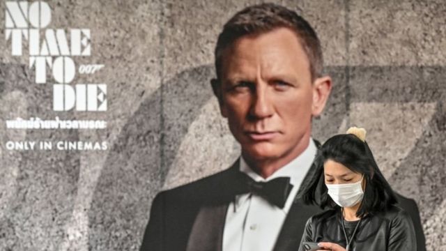 A woman wearing a face mask looks at her phone while standing in front of the new James Bond movie