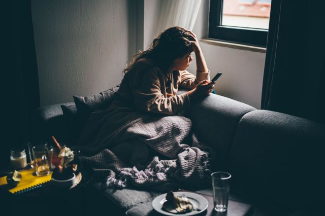 Una mujer mira por la ventana con un celular en la mano.