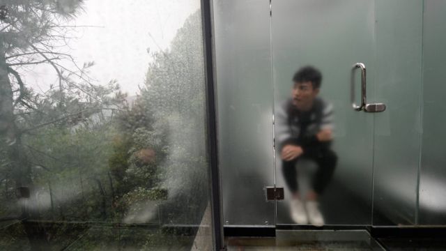 Hombre sentado en un inodoro detrás de una puerta de vidrio ligeramente esmerilado.