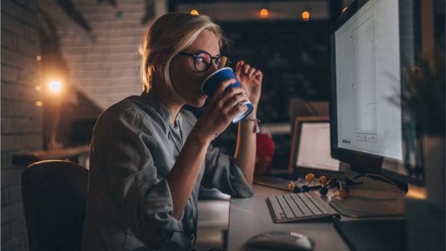 A woman drinking coffee from a paper cup and looking at a computer screen