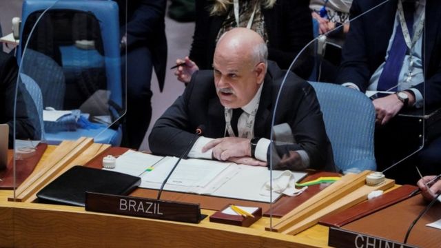 Embaixador discursando sentado, com placa escrita 'Brazil' à frente