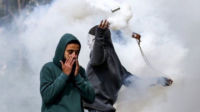 A Palestinian demonstrator covers his nose while another masked protester uses a slingshot to hurl back Israeli-fired tear gas cannister