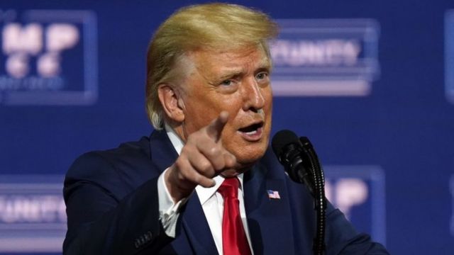 Trump falando ao microfone durante um evento eleitoral, dedo levantado