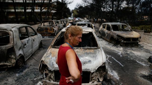 Veículos completamente destruídos pelo fogo na Grécia - em primeiro plano, uma mulher de blusa vermelha chora