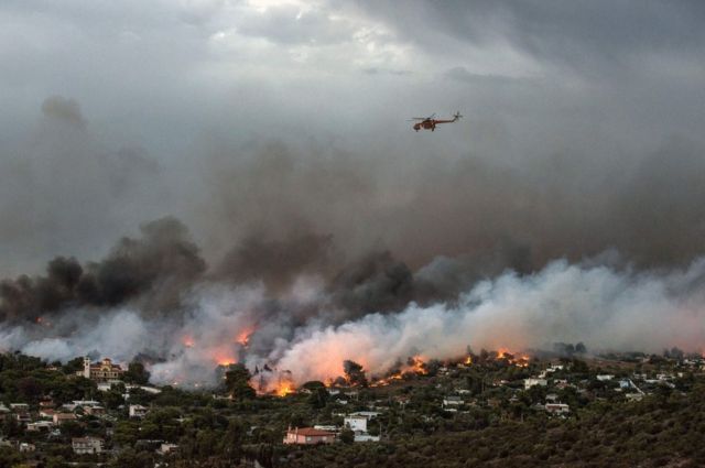 Imagem mostra helicóptero sobrevoando área com casas e vegetação onde podem ser vistas as chamas e a fumaça gerada pelo incêndio na Grécia