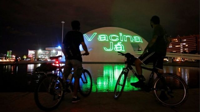 Duas pessoas em bicicletas diante de neon em que se lê vacina já