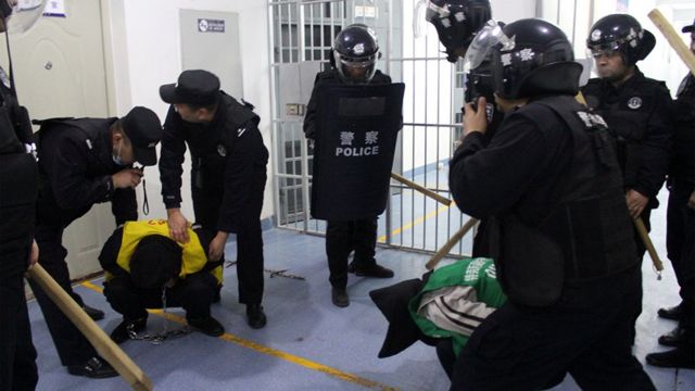 一名犯人蹲下，低着头，被手持棍棒的警官包围(photo:BBC)