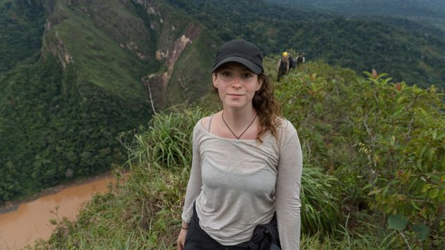 A diretora Elizabeth Unger, uma mulher branca jovem, em trilha em meio a montanhas cobertas por floresta