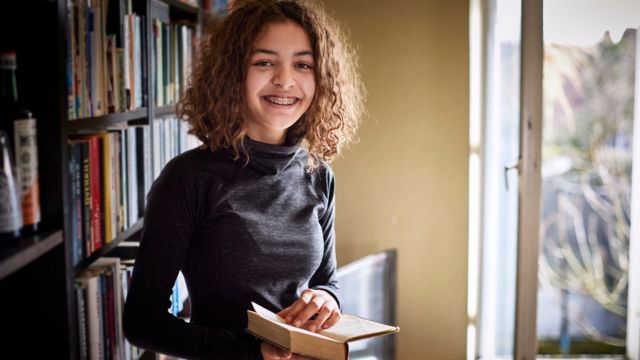 Jovem parda de cabelo cacheada segura livro na mão; uma estante e uma janela aberta ao fundo