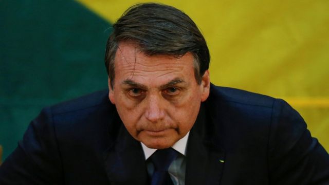 Bolsonaro aparece com feição séria, com cores verde e amarela no pano de fundo