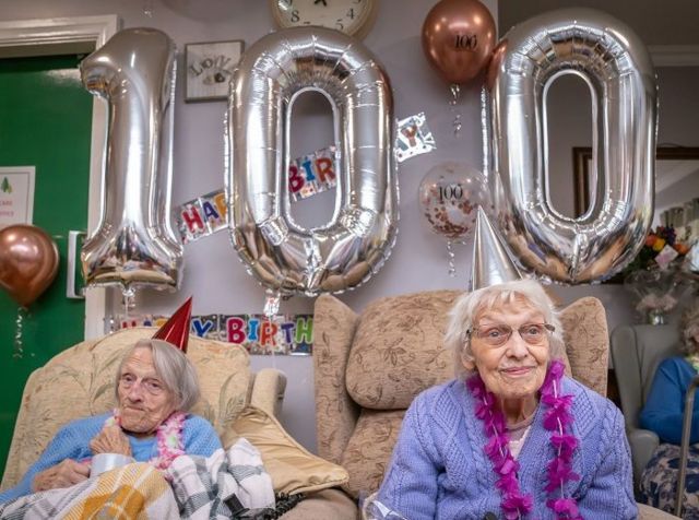 Duas senhoras sentadas em frente a balões em formato de 100