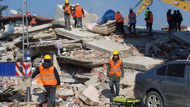 A team of rescuers, dressed in orange vests and helmets, walk through a pile of rubble.