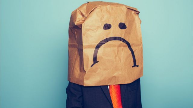 Imagen de alguien vistiendo un traje con corbata, pero que lleva una bolsa de papel en su cabeza con el dibujo de un rostro triste.