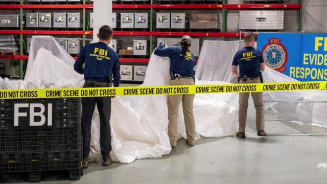 Funcionários do FBI mexem em grande objeto dentro de sala fechada, com faixa escrita em inglês: "Cena do crime, não ultrapassar"