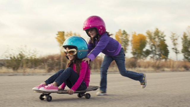 Two little girls are racing on skateboards, wearing helmets and smiling. One girl is pushing the other and they make a great team.